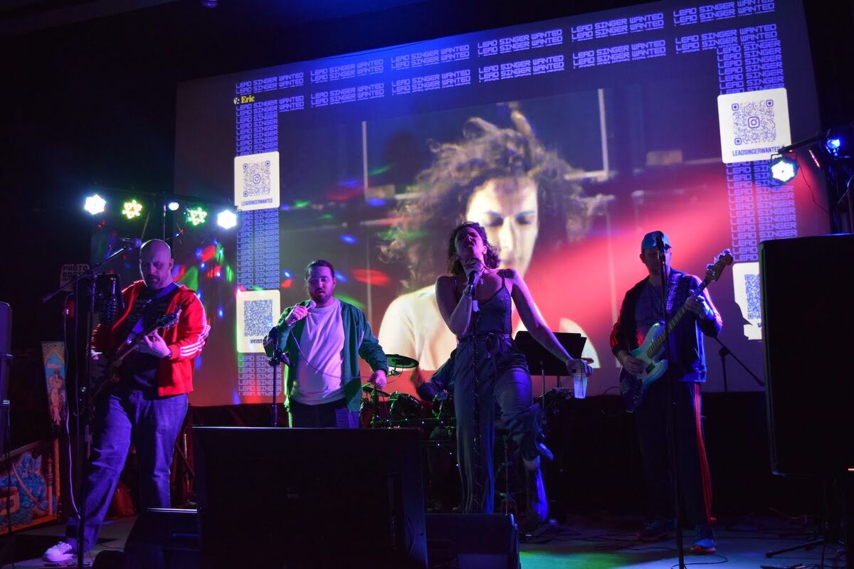 Singer performing with the full band — guitar, bass, drums — with colorful projected visuals and stage lights